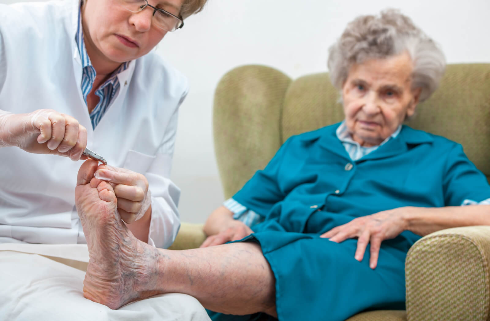 A female care professional is trimming the toenails of an elderly individual seated on a couch.