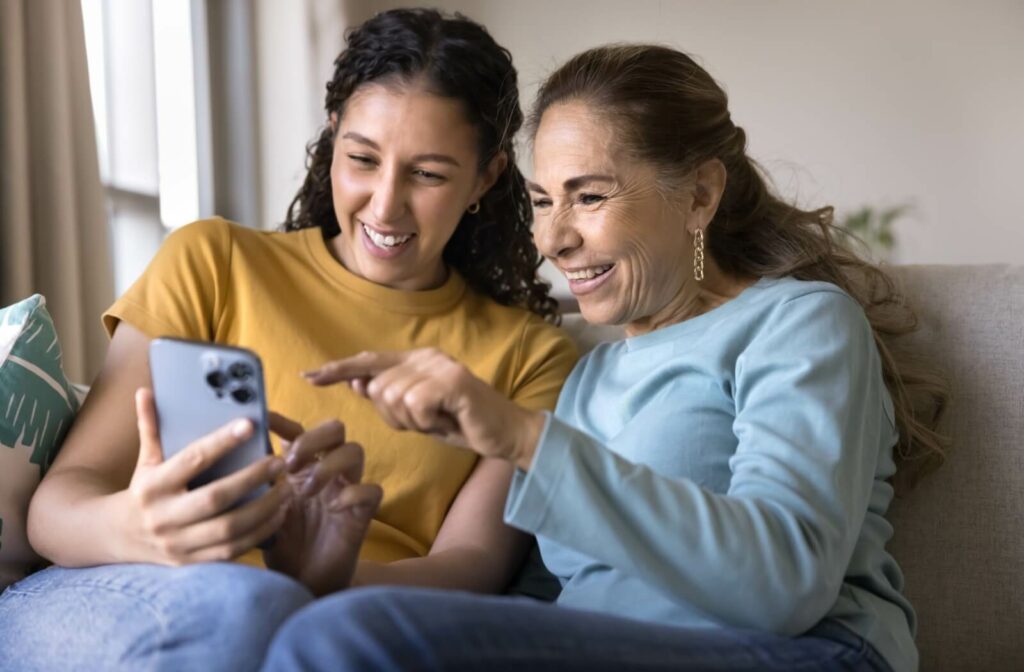 Older adult and younger adult sitting on couch smiling and pointing at smartphone while exploring remote learning together